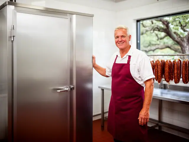 Smiling meat market owner in a red apron beside a new stainless-steel smokehouse cabinet